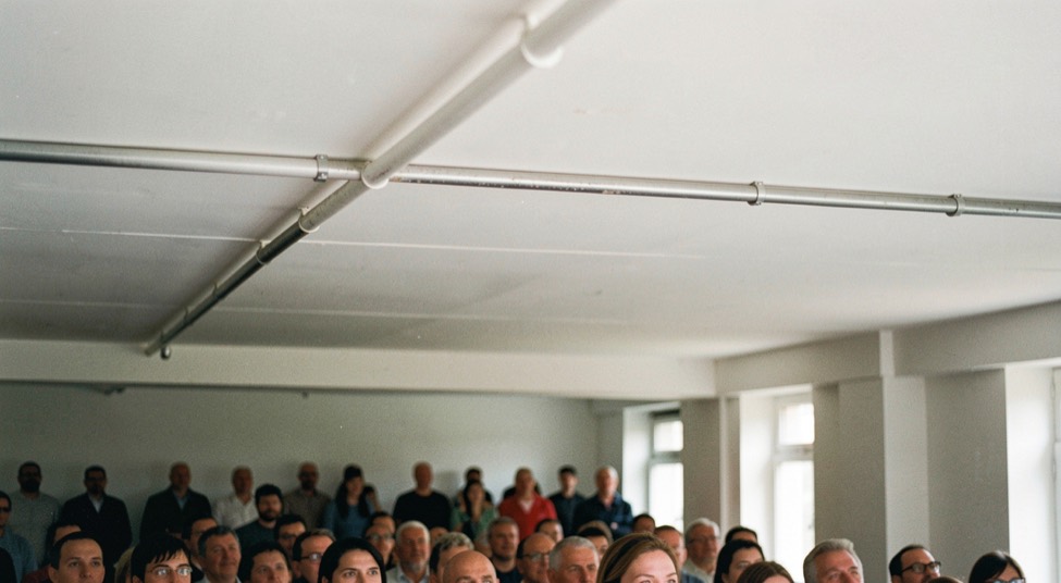 Low white ceiling above a small audience seen from the performer side, a heating pipe running across the top of the frame